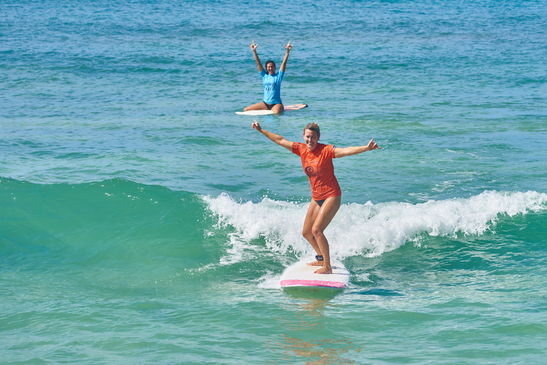 Two women riding a wave together on surfboards, arms raised in celebration